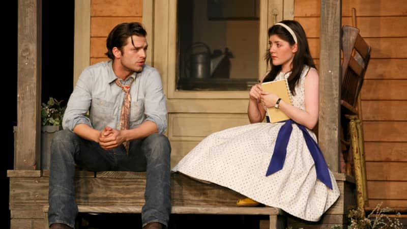 A man in a tattered denim skirt, old jeans, and a loose tie sits on porch steps and talks to a a young woman with a white dress and headband who holds a notebook.