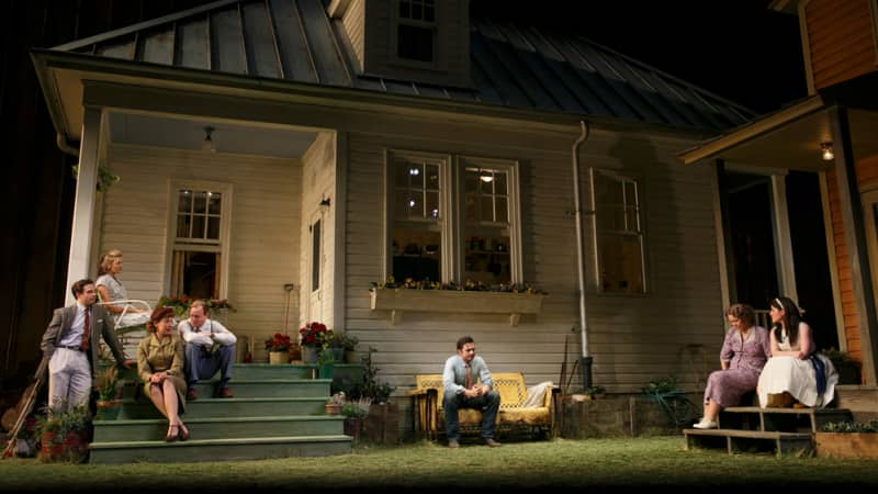 Seven actors in 1950s clothing sit on the back porch steps of two houses that share a backyard.