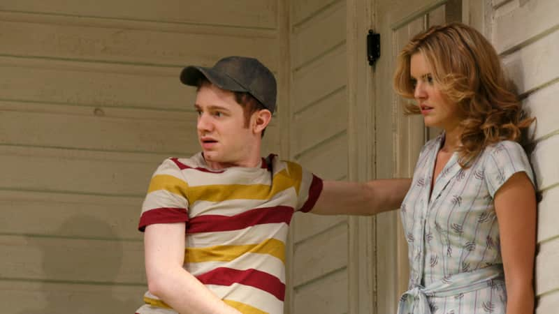 A young man and woman stand by the back door of a house. The woman presses her back against the wall, and the man holds an arm on the door.