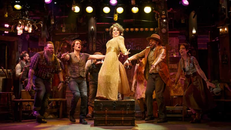 A woman in a gold dress dances on top of a trunk. People in southern clothing joyfull dance around her among yellow and purple lighting.