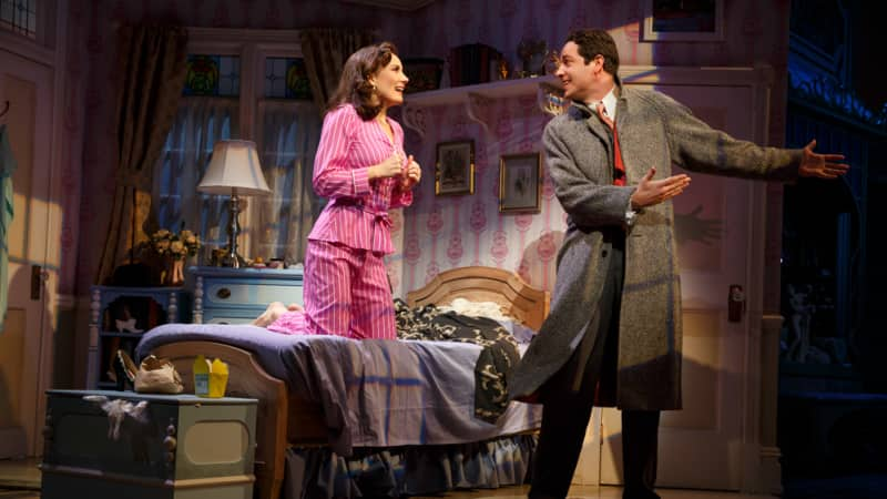 A woman in pink striped pajamas kneels on her bed while a man in a long coat gestures away from her. They both smile.