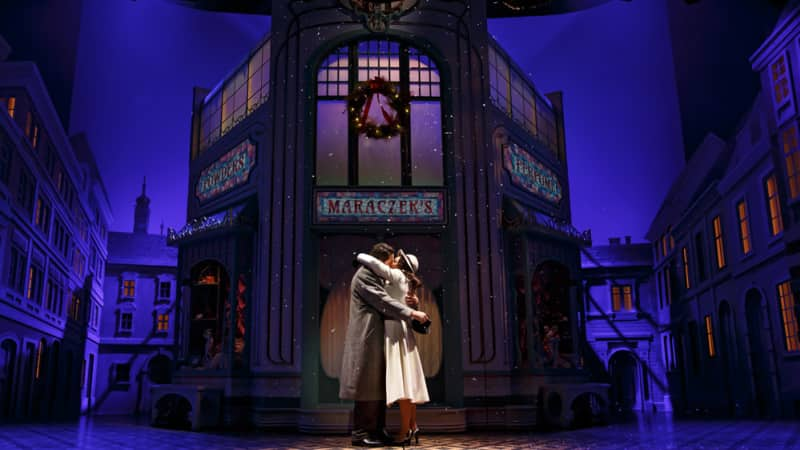 A spotlight shines on Zachary Levi and Laura Benanti kissing on stage in SHE LOVES ME. The set resembles a town sqaure.