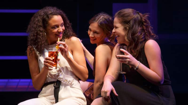 Three women in party outfits sit together on a step, laughing and sipping their drinks.