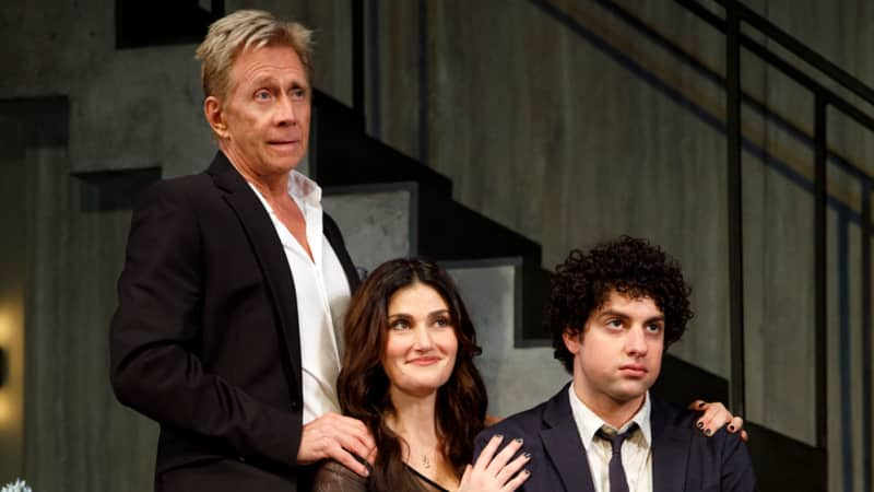 An older man, a woman, and a younger man pose for a family photo in a living room. The older man stands behind the woman and young man who sit on the couch.