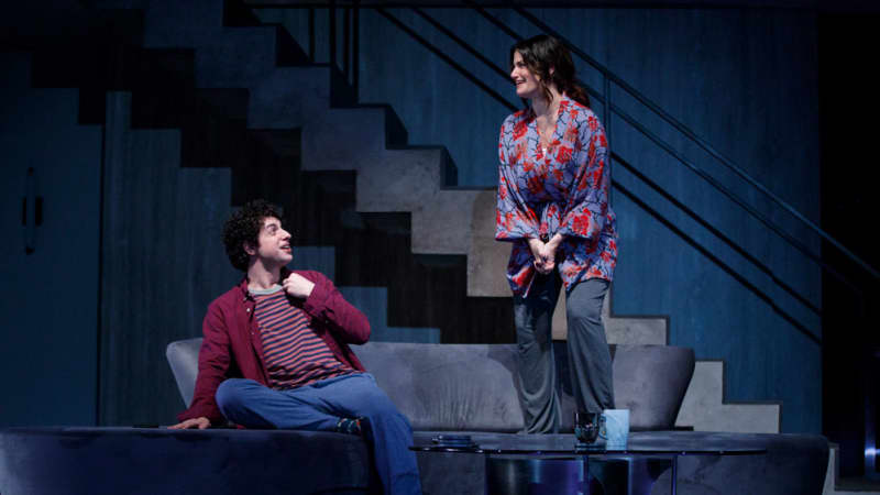 A woman in pajamas and a robe stands on a living room sofa. A young man in jeans and a striped shirt sits on the sofa and looks up at her smiling while leaning away.