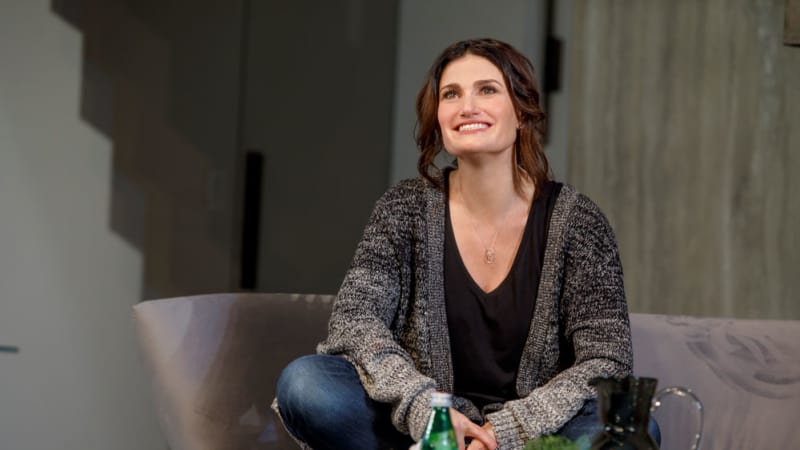 A smiling woman with long brown hair pulled into a loose ponytail sits on a couch with her legs crossed. On a table in front of her are snacks and sparkling water.