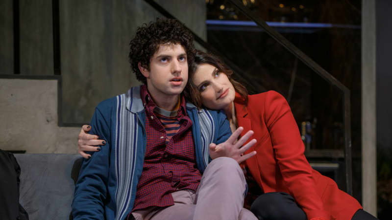 A woman in a red blazer sits on her knees on a sofa and hugs from the side a young man with curly dark brown hair who looks slightly annoyed.