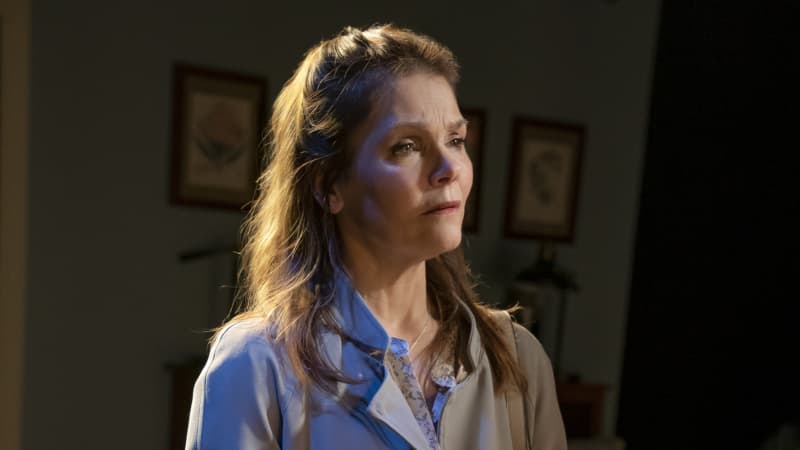 A sad, older white woman stares into the distance while she stands in a bedroom. She wears a coat, a purse, yellow rubber globes, and holds paperwork.