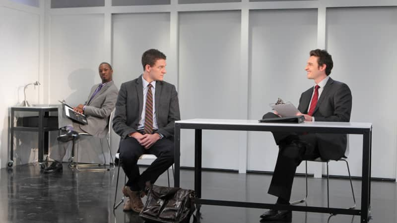 3 men in a room all wearing suits. It looks like an interview with one man smiliong and holding a clipboard. 