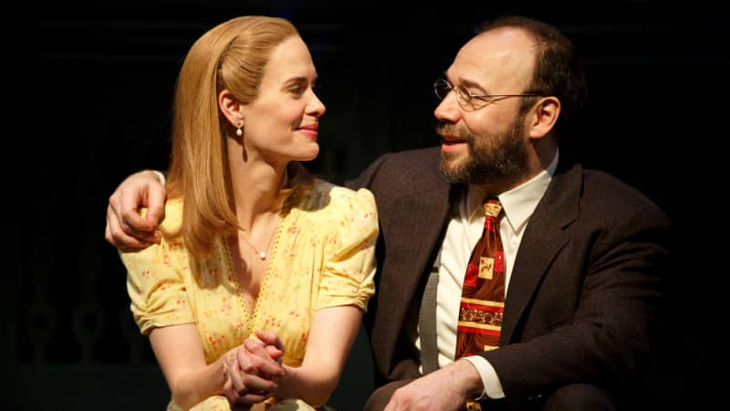 A man sits next to a woman and puts his arm around her as they both smile. She is blonde and wears a yellow 1940s dress, he has a beard and glasses and wears a suit.