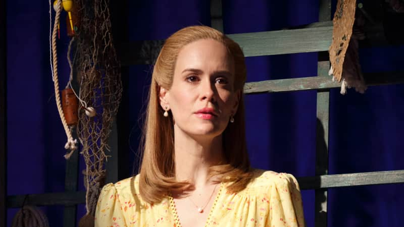 A woman with blonde hair wearing a 1940s yellow floral dress stares into the distance while holding a straw hat.