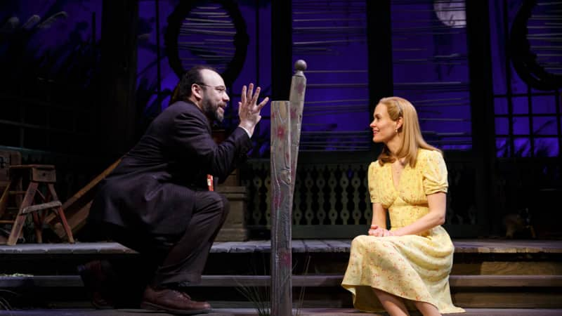 A man and woman sit together on wooden steps and have an engaging conversation, a railing separating them. He has glasses and a beard, she has blonde hair and wears a yellow 1940s dress.