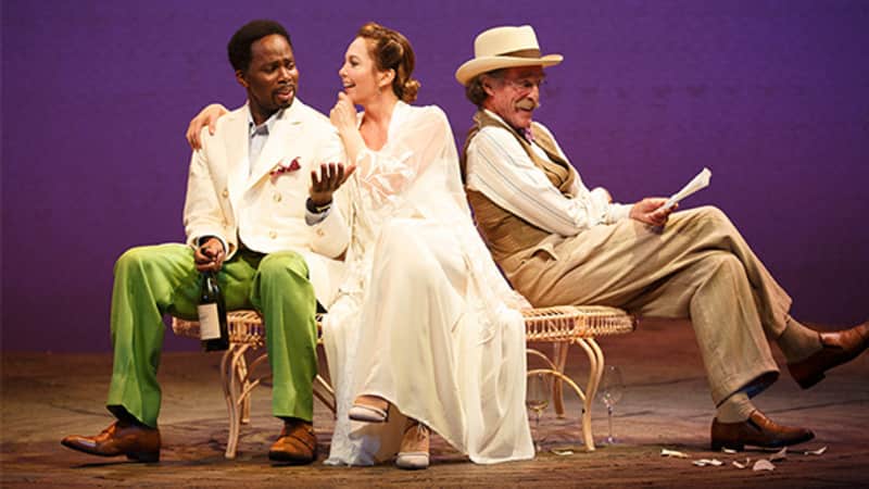 A woman in white sits on a garden bench between two men. One wears green pants, a white blazer, and holds a bottle of wine. The other wears a white hat and tan suit set while holding a paper.