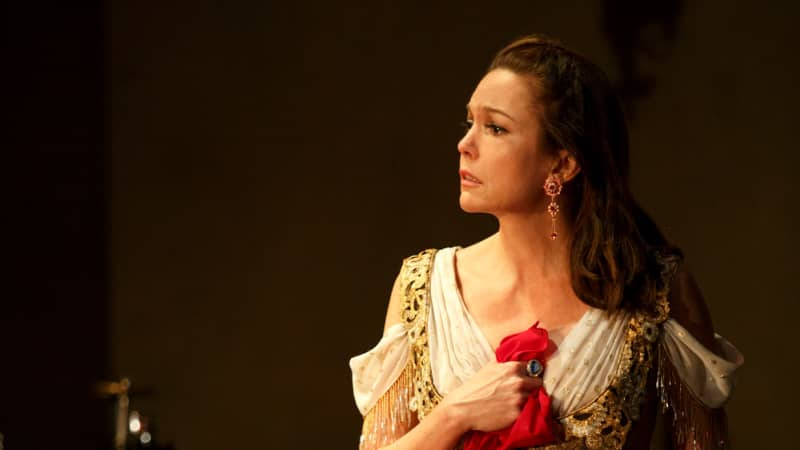 A woman in a golden corset and red patterned skirt sits on a chair and holds a handkerchief to her chest with sorrow.