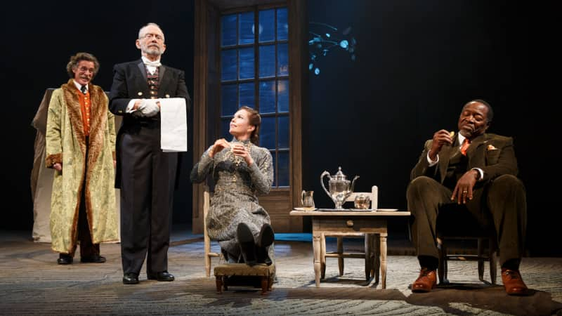 Two fancy people sit in small chairs and have tea at a small table. A butler stands to the side with a towel on his arm. Behind him is a man with a large fure coat.