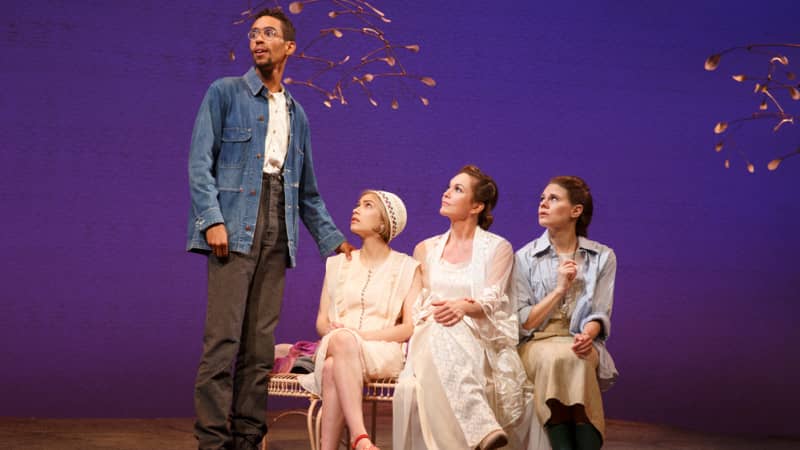 Three young woman in white sit on a garden bench and look up at a young man in glasses and a denim jacket, who places a hand on one of their shoulders. Wispy cherry trees are behind them.