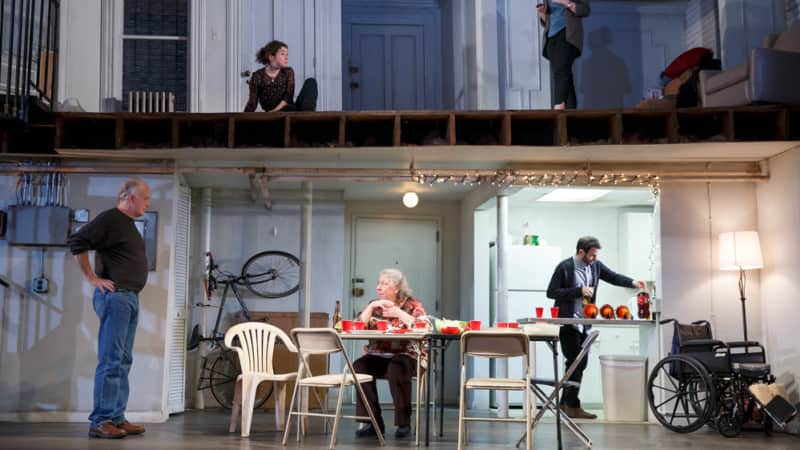 A split level set of a house. On the bottom in a basement, people sit and stand around a folding table with red Solo cups. On top are two women in a sparse room.