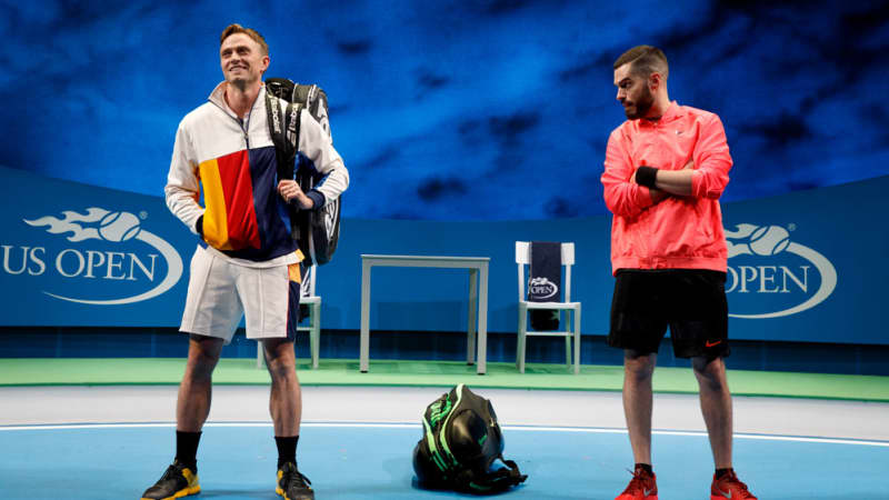 Two tennis players on the US Open tennis court. One of them slings his racket bag over his shoulder and smiles, while the other crosses his arms and glares at him.