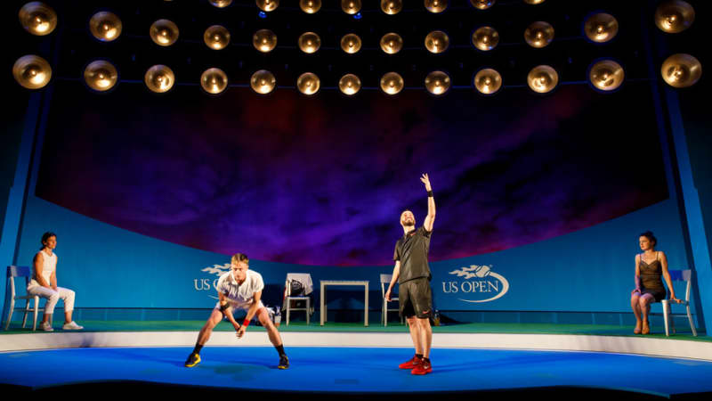 Two tennis players mime playing a tennis game on a large stage that looks like the US Open tennis court. A woman sits on a chair off to the side of the court.