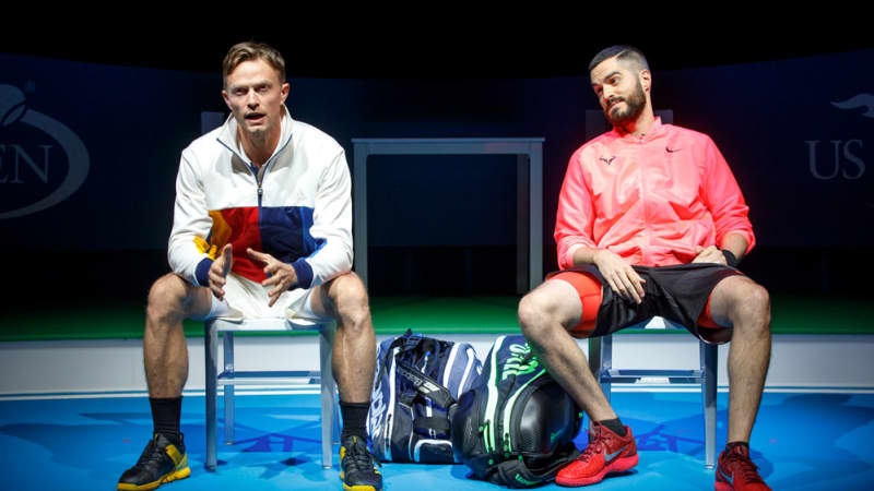 Two men in athletic clothes and zip up jackets sit on chairs on the side of a tennis court. Their gym bags sit between them.