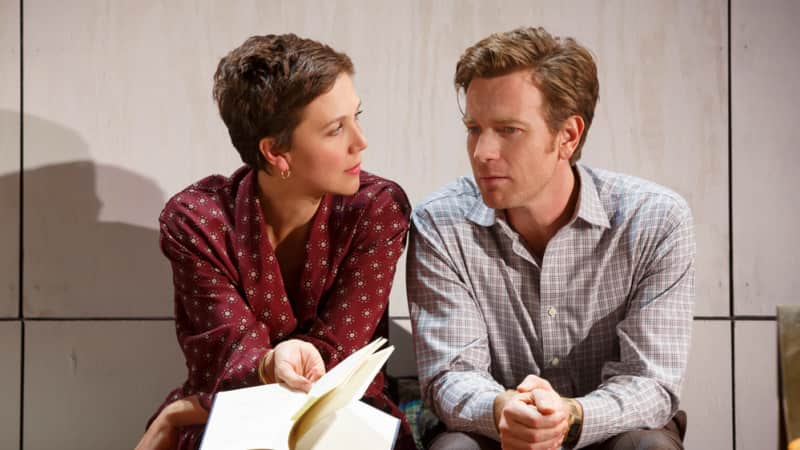 A woman with a brown pixie cut and a red dress sits down next to a man with reddish brown hair wearing a flannel. She holds a notebook in her hand.