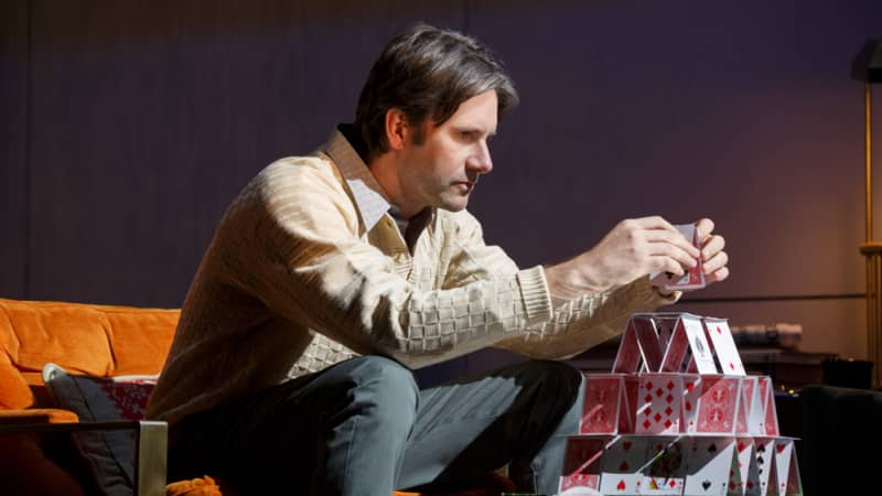 A man in a light yellow sweater and white collared shirt builds a house on a coffee table using playing cards. He sits on an organe sofa.