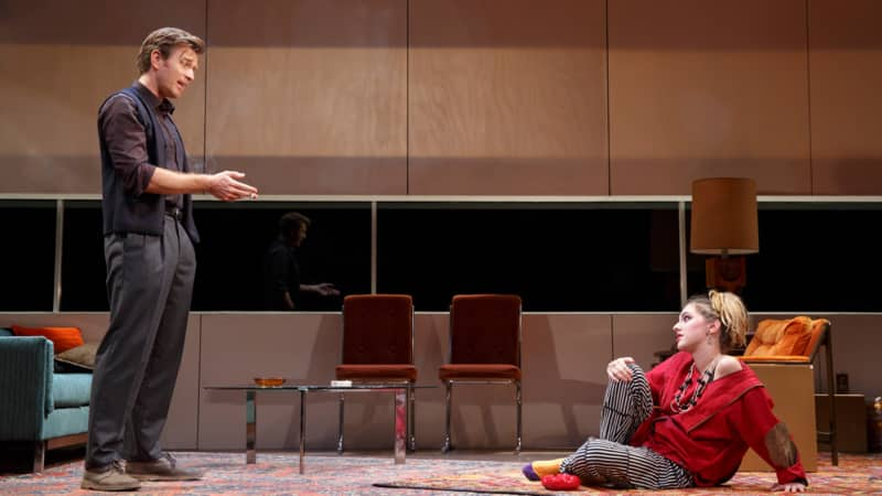 A man in a knit vest and collared shirt stands and speaks down to a woman in red and black lounging on the floor with a hand on her knee.