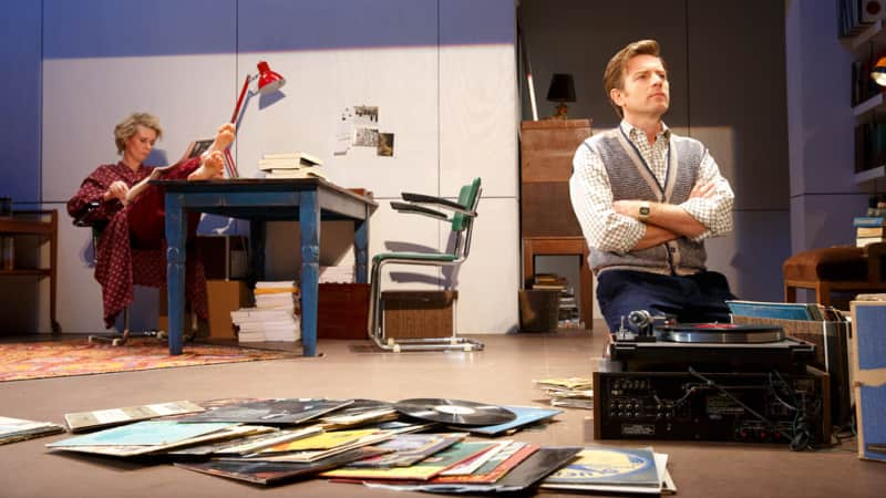 A man kneels on the floor in front of a record player and several vinyls on the floor. Behind him, a woman reads from a magazine with her feet propped up on a desk.