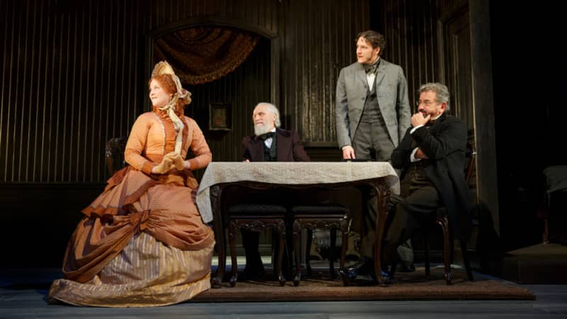 A group of upperclass 1860s-styled people sit and stand around a small table in a drawing room. Three men of varying ages in suits. One woman in an intricate orange dress and bonnet.