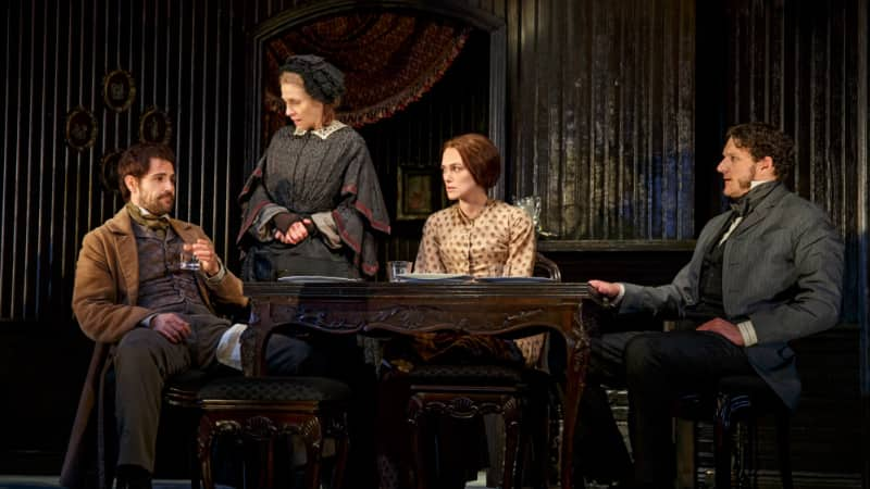 Four actors in 1860s style clothing sit together at a dark wooden table and have a tense interaction. They all look at one man who drinks from a glass.