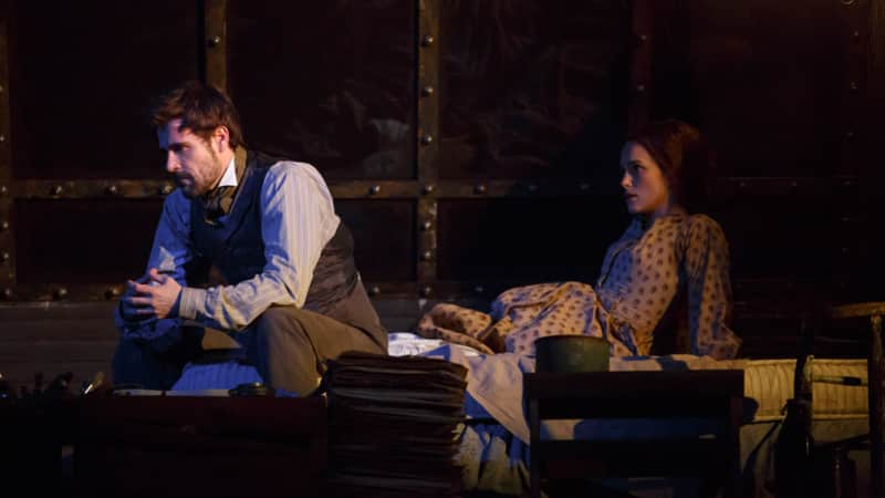 A man in 1860s suit pieces sits on the end of a bed, resting his elbows on his knees. A woman in a brown patterned dress sits up in bed behind him. They both wear serious expressions.