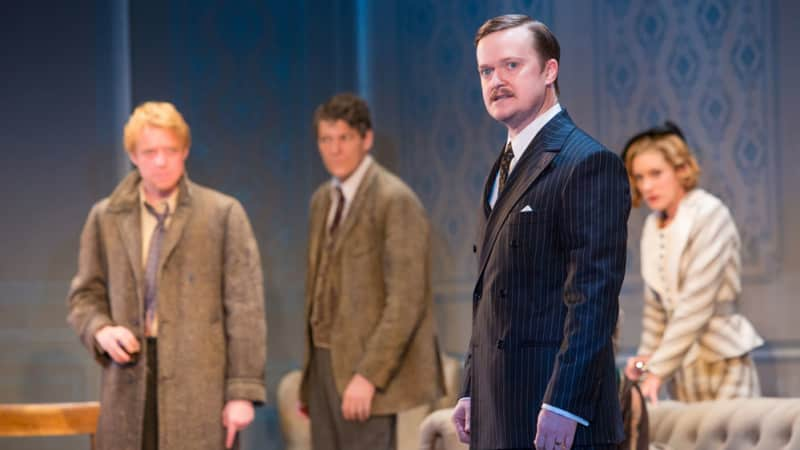 A man with slicked hair and a small mustache in a pinstriped suit looks lost in thought. Behind him several people stare at him with tentative expressions. 