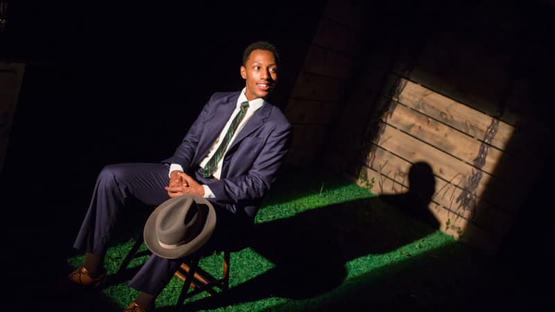 A rectangular spotlight on a Black man in a suit resting in lawn chair, a fedora balancing on his knee. His shadow is sharply cast behind him, and the rest of the room is pitch black.