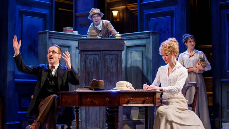 A man and woman in 1910s clothing sit together at a wooden desk, their hats resting on the table, and books piled below them. An older man stands at podium behind them and a woman holds some books.