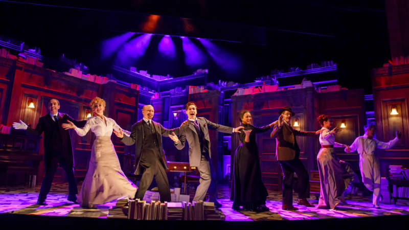 A line of actors in 1910s clothing dance together at the front of a stage. Pink and purple lighting bounces across the set, which looks like an old study.