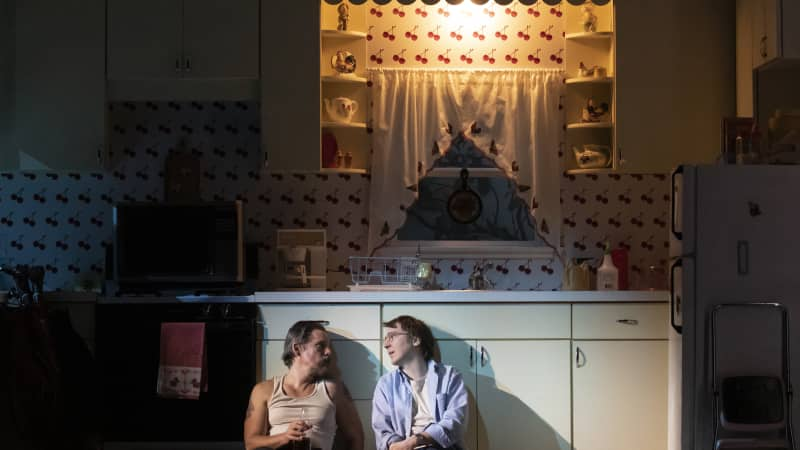 Two men sit on the floor of a kitchen set with their backs against the cabinets. The room is only lit by one light above the sink. One of them holds a beer bottle.