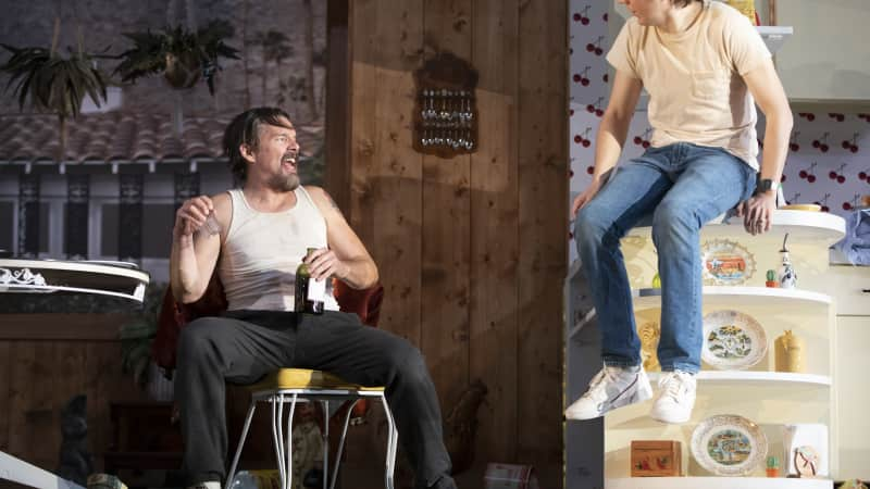 A man in a white tank top holding a beer bottle laughs and sits in a chair in a messy home, with beer cans scattered around. Another man sits on the kitchen counter and looks at him.