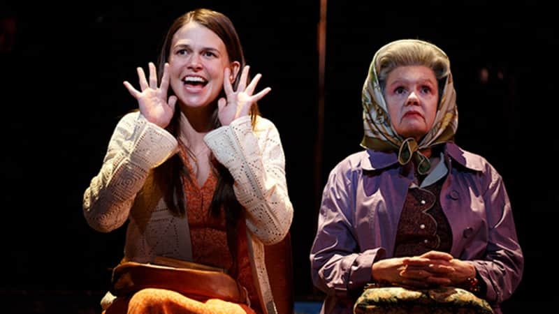 A woman with long brown hair wearing an orange dress holds her hands up on the sides of her cheeks like she's shouting. An old woman wearing a hair scarf sits next to her.