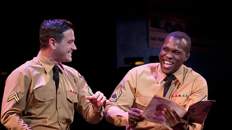 Two men in tan military uniforms hold magazines and laugh with each other.