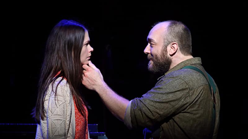 A man with a beard reaches an arm out and holds the chin of a woman with long brown hair wearing an orange dress and white cardigan.