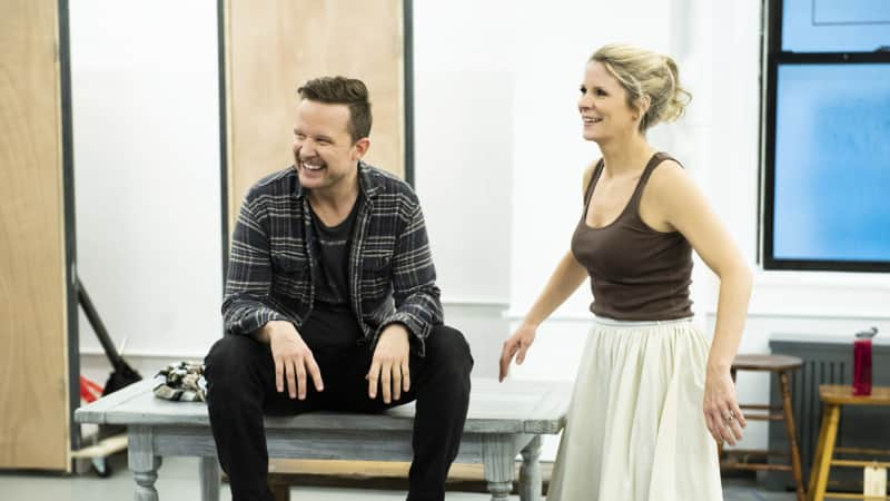 A man sits on a table in the rehearsal studio and a woman stands next to him while they both smile and laugh.