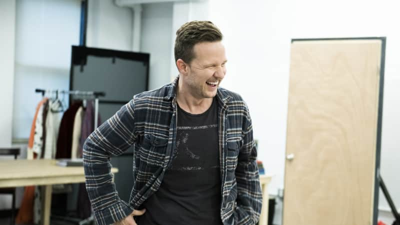 A man laughs in the rehearsal studio. He wears a black flannel over a black t-shirt and jeans.