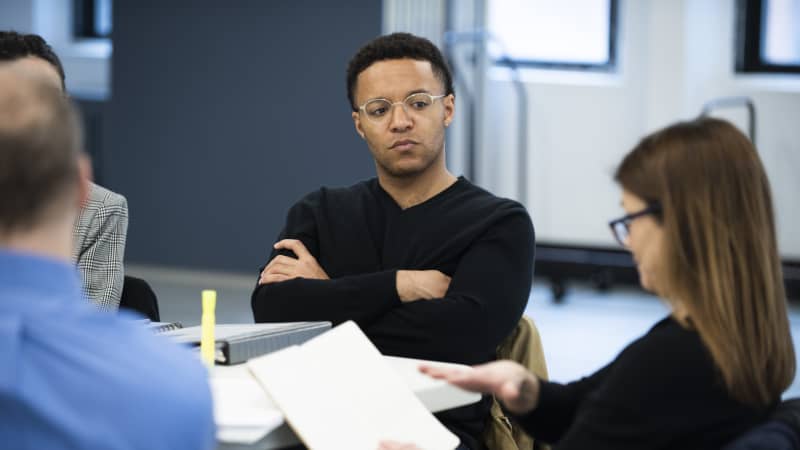 A young Black man wearing round silver glasses crosses his arms and listens to the table read.