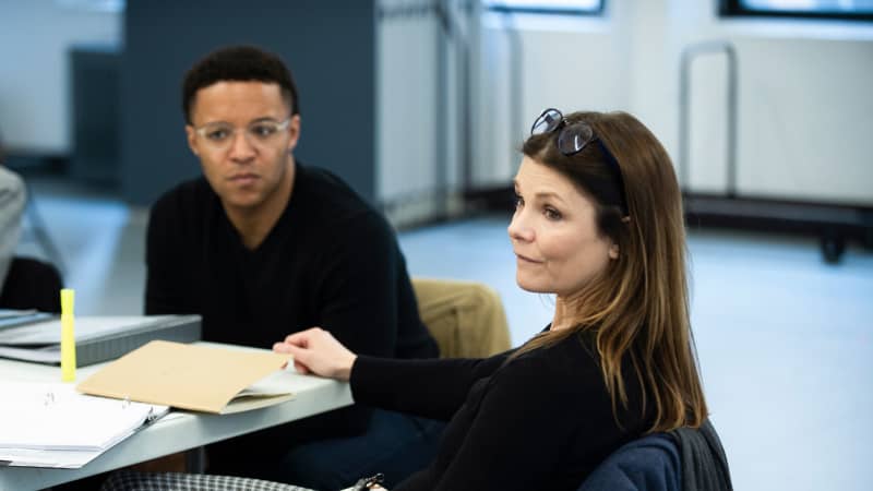 A young Black man and an older white woman sit next to each other at a rehearsal table and listen intently.