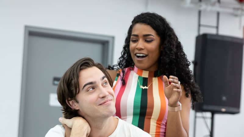 A young woman puts her arm around a young man who sits sideways on a wooden chair in the rehearsal studio.