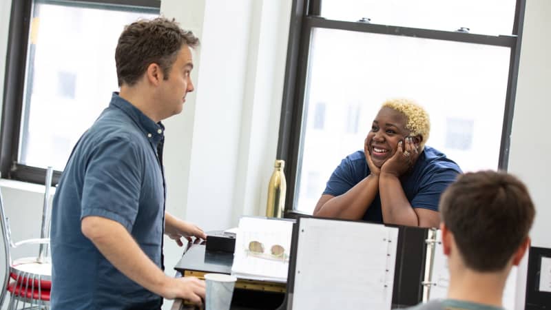 A woman rests her hands on her cheeks and smiles while she leans on a black piano and speaks with a man standing at the piano.