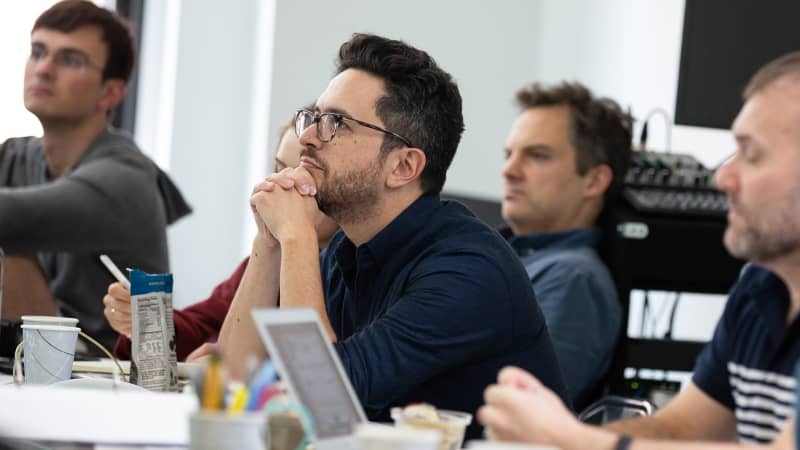 A man with glasses sits behind a fold out table with other creative team members to watch rehearsal.