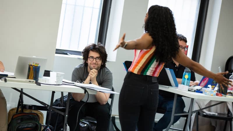 A man with long hair and round glasses smils while watching an actress sing with her arms outstretched in the rehearsal studio.