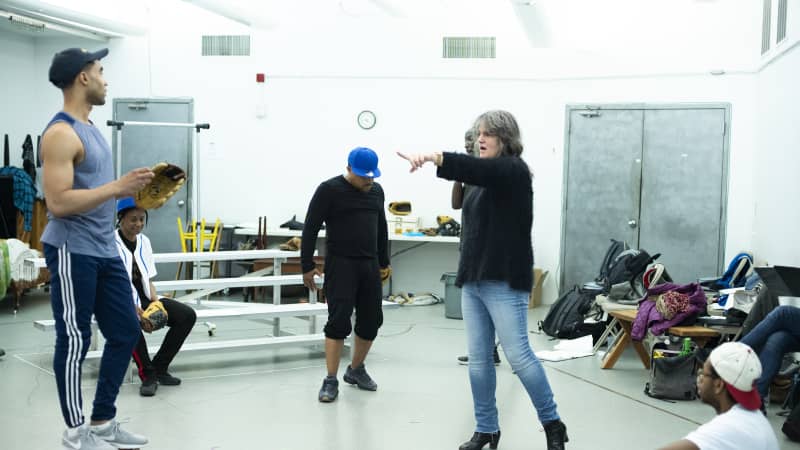 A woman points to actors dressed as baseball players who stand in the rehearsal studio, one sitting on metal bleachers.