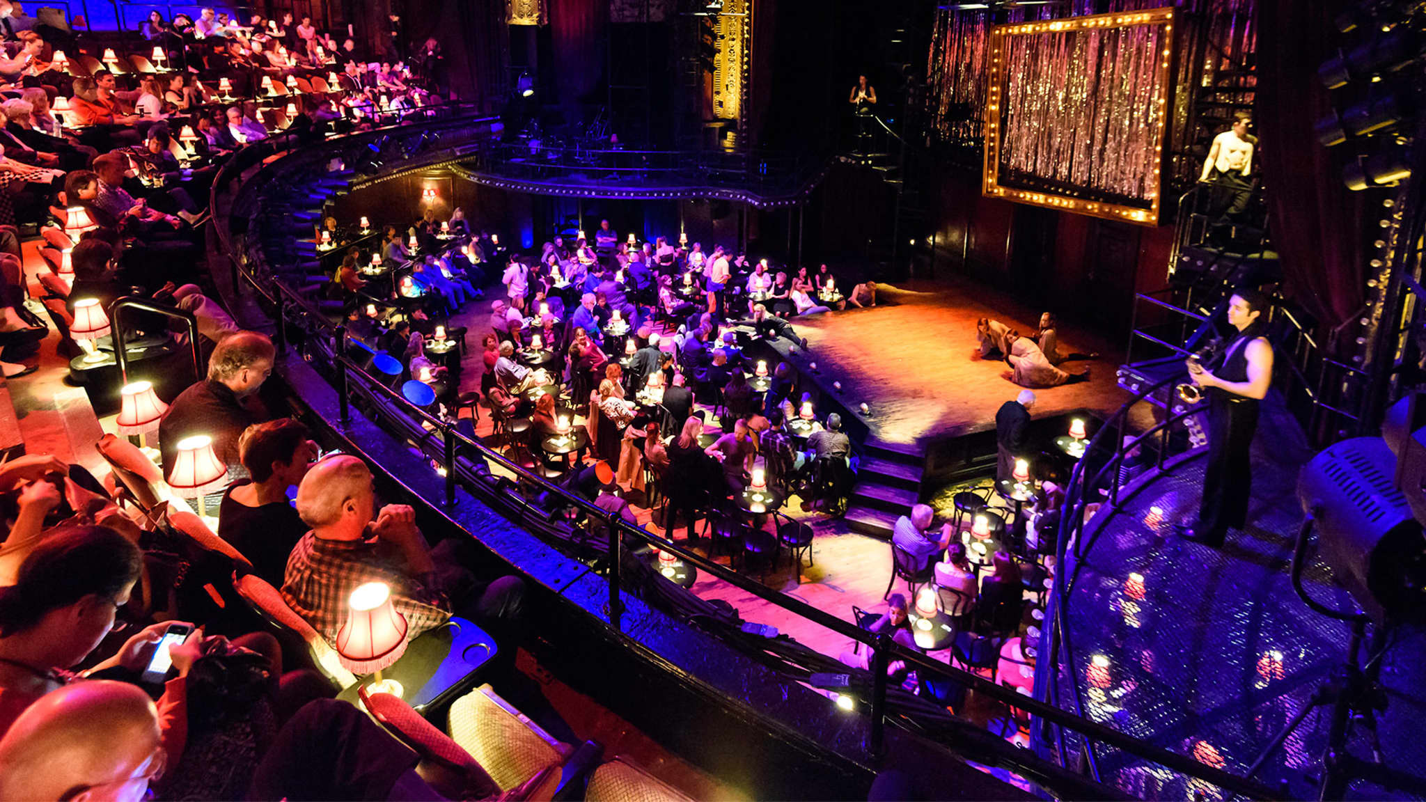 The audience at Studio 54 during CABARET. The orchestra level is full of small tables, chairs, and lamps. Orange, purple, and blue light shines around the theatre.